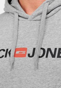 Grå hættetrøje med snøre, med sort "JONES" tekst og en rød firkantet detalje med et indlejret logo. Lavet af blødt stof.
