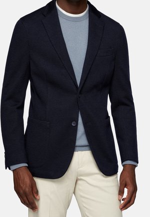 Mężczyzna w ciemnoniebieskiej marynarce, jasnoniebieskim swetrze i kremowych spodniach, stojący z rękami swobodnie opuszczonymi wzdłuż ciała.