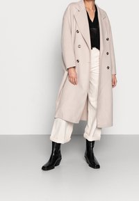 Manteau croisé beige clair avec de gros boutons, associé à un haut noir, un pantalon crème à jambes larges et des bottines à talons noires.