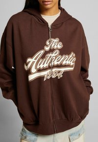 Hnědá mikina s kapucí na zip se velkým bílým potiskem "The Authentic 1994" na přední straně. Obsahuje nastavitelnou kapuci a vroubkované manžety.
