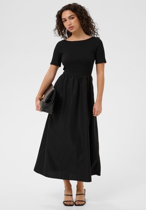 Vrouw met krullend haar die een zwarte maxi-jurk met korte mouwen draagt, een grote zwarte clutch vasthoudt en zwarte bandjeshakken draagt.