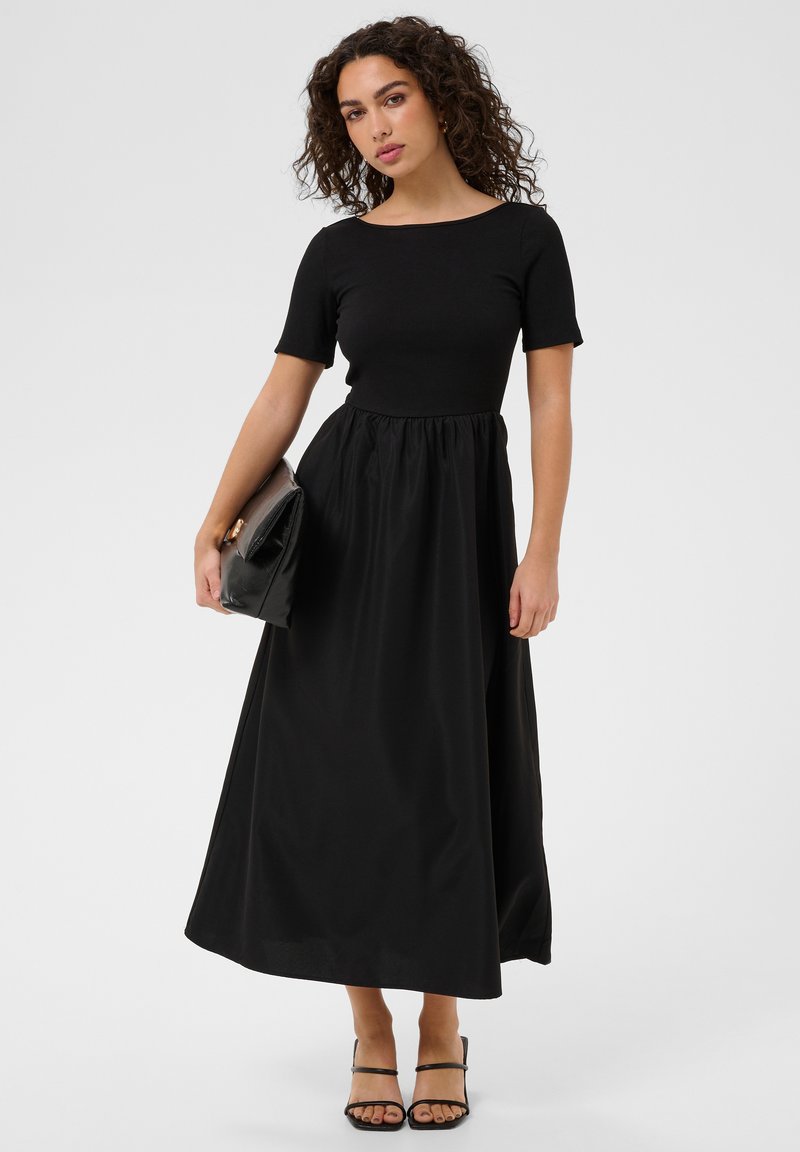 Femme aux cheveux bouclés portant une robe maxi noire à manches courtes, tenant une grande pochette noire et portant des talons noirs à brides.