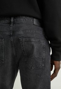 Hombre que lleva jeans de mezclilla negros desgastados y una camiseta de manga larga negra, mostrado de la cintura a la mitad del muslo.