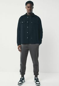 Black button-up shirt with two chest pockets, paired with gray joggers and black sneakers featuring white accents. Smooth, casual design.