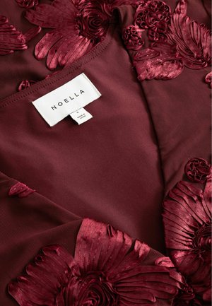Burgundy tyg med tredimensionell, intrikat mönstrad blommig broderi. Tagg synlig med varumärket "NOELLA" och storleken "S."