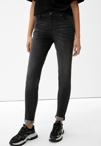 Jeans ajustados negros de tiro alto con un ligero desgaste, cinco bolsillos y dobladillo enrollado, combinados con zapatillas negras y grises.