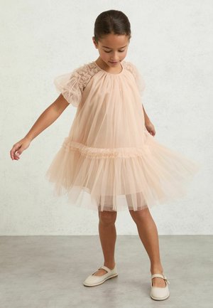 Fată tânără purtând o rochie ușoară din tul de culoare piersică deschisă, cu mâneci și fustă cu volane, privind în jos în timp ce ține rochia desfăcută în lateral.