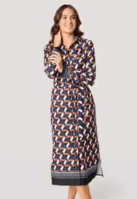 Lange, gemusterde jurk in marineblauw, met geometrische vormen in oranje, wit en zwart. Bevat een knoopsluiting aan de voorkant en een riem.