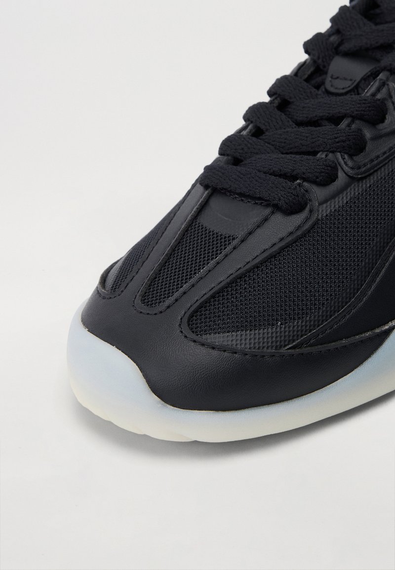Baskets noires présentant une combinaison de cuir texturé et de mesh. Le design comprend un bout rond et une semelle en caoutchouc translucide épaisse.