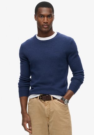 Man wearing a navy blue crewneck sweater over a white shirt, beige pants with a brown belt, and a black wristwatch on left wrist.