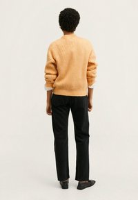 Gebreide oranje trui met geribbelde textuur, gecombineerd met zwarte rechte broek en zwarte platte schoenen. Witte manchetten zichtbaar aan de mouwen.