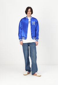 Jachetă bomber din satin albastru cu un logo alb "H", cu manșete dungi ribbate, asociată cu un tricou alb și blugi largi albaștri. Complectată cu adidași.