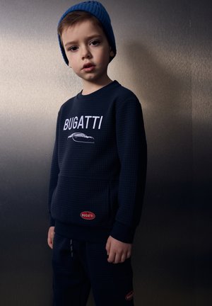 Giovane ragazzo con felpa Bugatti blu navy e pantaloni coordinati e un berretto di lana blu, in piedi davanti a un muro metallico scuro.