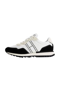 Zapatilla deportiva blanca y negra con parte superior de malla, acentos de ante texturizado, rayas contrastantes y suela de goma negra diseñada para tracción.