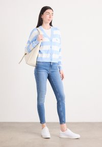 Cardigan rayé bleu clair et blanc avec un col en V, fermeture à boutons et logo brodé, porté sur une chemise blanche avec un jeans bleu et des baskets blanches.