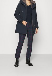 Marinblå parka med en huva fodrad med fuskpäls, kombinerad med rutiga byxor i mörkgrön, lila och blått, kompletterat med svarta ankelskor.