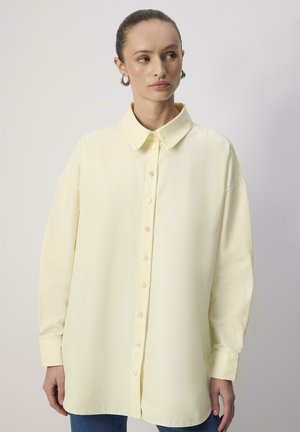 Femme aux cheveux attachés, portant une chemise ample jaune pâle à boutons et des boucles d'oreilles créoles, regardant sur le côté devant un fond uni.