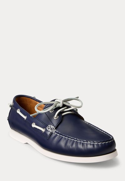 Scarpa da barca in pelle blu navy con cuciture bianche, lacci bianchi annodati a fiocco e suola in gomma bianca.