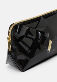 Funda de charol negro con un gran lazo decorativo, cremallera dorada y una placa de logo dorada que presenta "Ted Baker London". Textura suave y brillante.