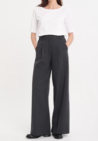 Höga, vida byxor i mörkgrått tyg med framfickor och veck, som bärs tillsammans med en figurnära vit T-shirt.