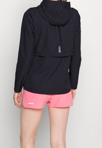 Schwarze Jacke mit Kapuze, aus glattem Material, hat eine taillierte Passform und reflektierende Akzente. Kombiniert mit pinken Shorts, die ein sportliches Design zeigen.