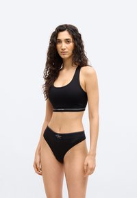 Νεαρή γυναίκα με μακριά σγουρά μαλλιά, φοράει μαύρο αθλητικό σουτιέν Calvin Klein και ταιριαστό εσώρουχο με ψηλό κόψιμο, στέκεται μπροστά σε ένα απλό φωτεινό φόντο.