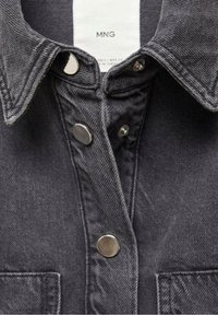 Jeanshemd in Dunkelgrau, mit spitzem Kragen, metallischen Druckknöpfen und zwei Brusttaschen mit sichtbarer Steppnaht.