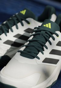 Baskets blanches avec des accents vert foncé, une tige texturée, des rayures noires et une semelle en caoutchouc robuste. Les lacets sont de couleur vert foncé.