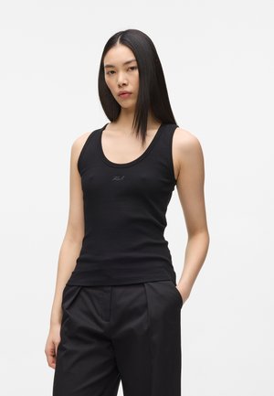 Jeune femme aux longs cheveux noirs portant un débardeur noir ajusté et un pantalon noir ample, debout avec une main dans la poche.