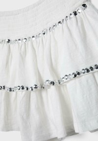 Jupe en tissu blanc froncé avec une ceinture élastique, décorée de deux rangées horizontales de sequins argentés.