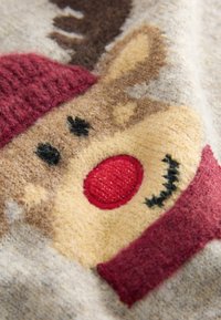 Gros plan sur un tissu doux avec un visage de renne tricoté, présentant un nez rouge, des yeux noirs et des cache-oreilles rouges sur un fond beige.
