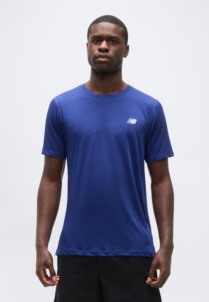 Νεαρός άνδρας που φοράει απλό μπλε κοντομάνικο μπλουζάκι New Balance και μαύρα σορτς, στέκεται μπροστά σε απλό φωτεινό φόντο.