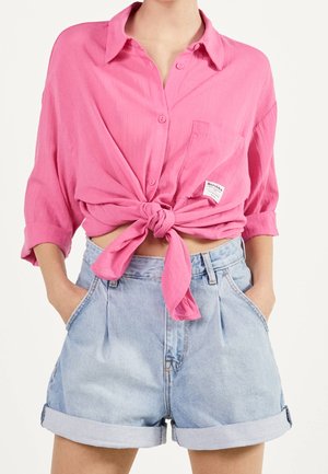 Vrouw draagt een roze overhemd met knopen, gestrikt in de taille, met opgerolde mouwen, en lichtblauwe, hooggetailleerde spijkershorts met opgerolde zomen.