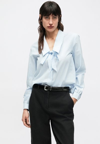 Blusa azul claro con rayas, gran cuello y detalle de lazo, combinada con pantalones negros y cinturón negro. Presenta mangas largas y un corte holgado.