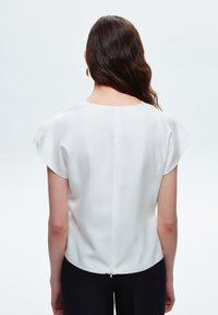 Blusa bianca a maniche corte in tessuto leggero con scollatura rotonda, zip sul retro e texture liscia. Indossata con pantaloni neri.