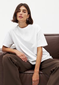 Hvid bomulds t-shirt med rund hals og korte ærmer, parret med løse brune bukser, siddende på en brun lædersofa.
