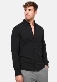 Camisa negra de manga larga con botones y cuello, con una textura suave y un corte slim. Con pliegue de botones horizontal y un ligero detalle en el pecho.