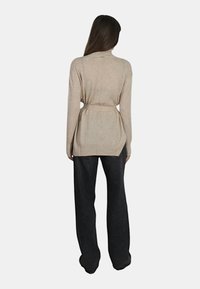 Cardigan in maglia beige con vita aderente, maniche lunghe e schiena leggermente aperta. Indossato sopra pantaloni neri a gamba larga.