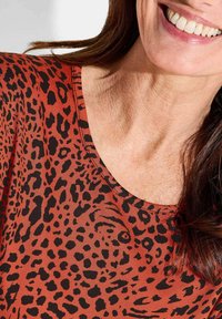 Top met luipaardpatroon en een ronde halslijn, met een roest-oranje achtergrond en zwarte vlekken, gemaakt van zachte stof. Glad textuur.