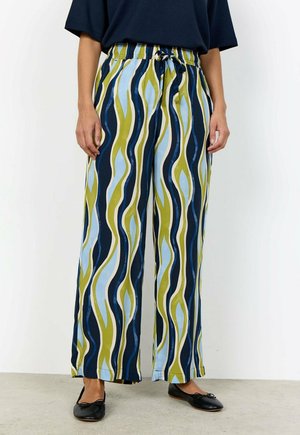 Persoon die wijde broek met blauw, groen en wit golvende strepen draagt, een donker bovenstuk en zwarte ballerina's, staande op een grijze vloer.