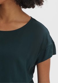 Tmavě zelený top s krátkými rukávy a kulatým výstřihem, s volánkovými rukávy, vyrobený z hladké látky, s volnějším střihem.