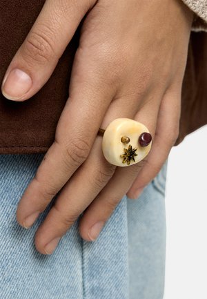 Dłoń z gołymi paznokciami nosząca masywny pierścionek z kremowymi, złotymi i ciemnoczerwonymi elementami dekoracyjnymi na jasnoniebieskim denimu i brązowej tkaninie.