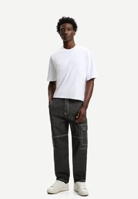 Witte t-shirt met korte mouwen, gecombineerd met donkergrijze cargobroek met witte stiksels en zijzakken. Gedragen met witte sneakers.