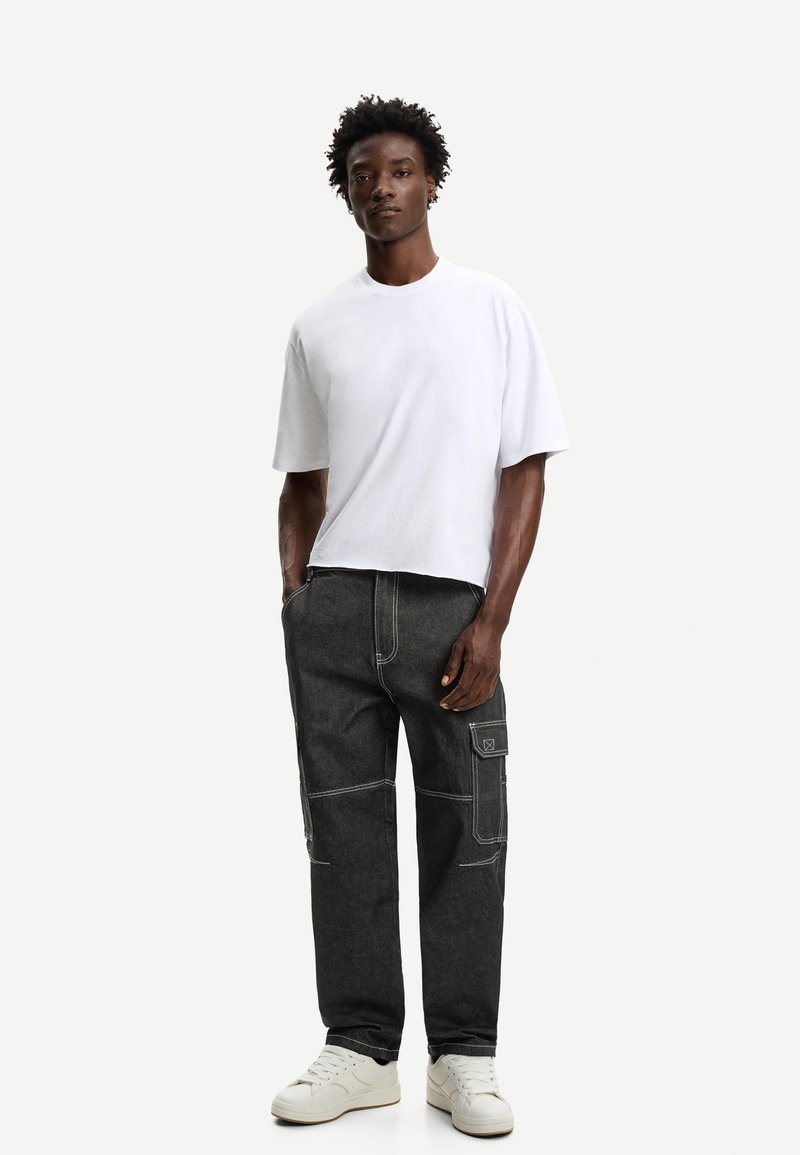 Witte t-shirt met korte mouwen, gecombineerd met donkergrijze cargobroek met witte stiksels en zijzakken. Gedragen met witte sneakers.