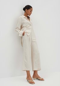 Beige, knoopshirt met brede pijpen, lichtbeige broek en beige slippers. Heeft een ontspannen pasvorm en een gladde textuur.