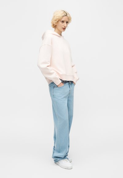 Άτομο με κοντά ξανθά μαλλιά, φοράει ανοιχτό ροζ φούτερ με κουκούλα, φαρδιά ανοιχτό μπλε τζιν και λευκά αθλητικά παπούτσια, στέκεται με τα χέρια στις τσέπες.