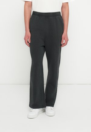 Άτομο που φοράει μαύρη φαρδιά φόρμα με ελαστική μέση και λευκά αθλητικά, στέκεται μπροστά σε απλό λευκό φόντο.