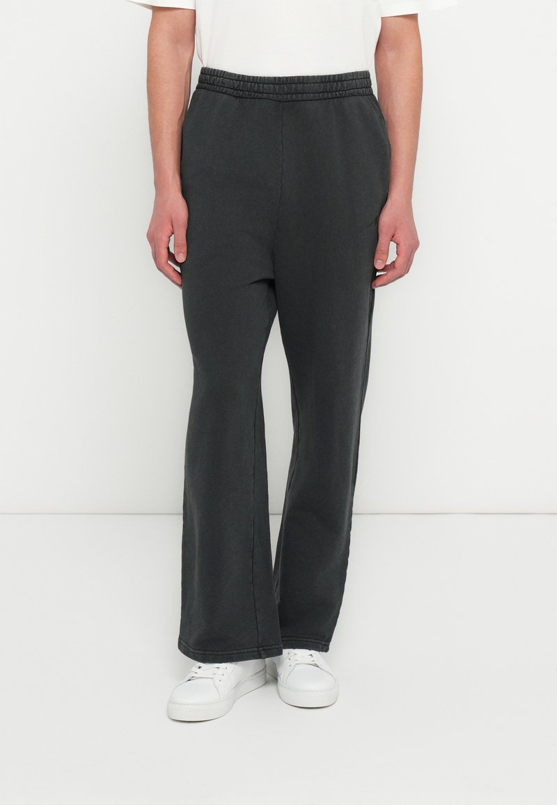 Άτομο που φοράει μαύρη φαρδιά φόρμα με ελαστική μέση και λευκά αθλητικά, στέκεται μπροστά σε απλό λευκό φόντο.