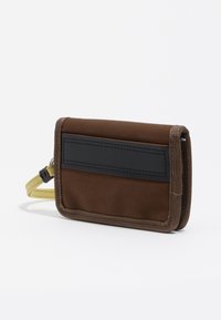 Cartera de tela marrón con bordes acolchados, parche negro cosido en la parte frontal y correa amarilla para la muñeca unida a un lado.