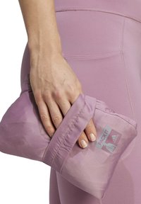 Leicht lila packbare Jacke in der Hand, aus leichtem Stoff mit elastischen Bündchen und einem Logo-Patch. Glatte Textur, kompaktes Design.